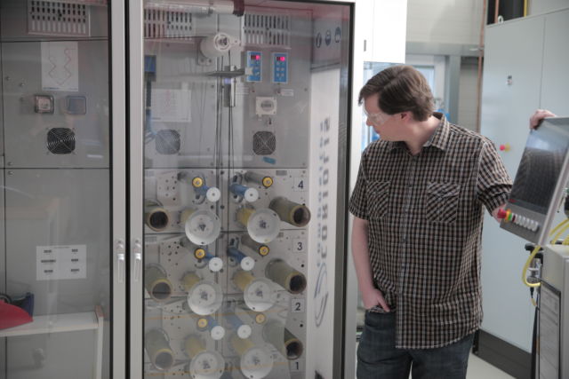 Ars' Lee Hutchinson stands in front of the creel cabinet that feeds carbon fiber to the robot that took all of our carbon fiber layup jobs.